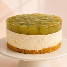 Load image into Gallery viewer, A round no-bake earl grey cheesecake with green grape and jello on top, displayed on a white cake stand against a warm beige background.
