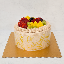 Load image into Gallery viewer, A round chocolate cake with piped cream borders, topped with fresh fruit including strawberries, blackberries, green grapes, and pineapple, presented on a gold cake board against a light marble background.
