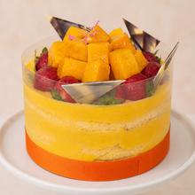Load image into Gallery viewer, A round mango mouuse cake with fresh yellow mango and strawberry toppings, displayed on a white cake stand against a light beige background.
