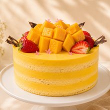 Load image into Gallery viewer, A yellow layered mango mousse cake with fresh mango cubes, strawberries, and chocolate shavings on top, displayed on a white cake stand against a warm beige background with soft floral props.
