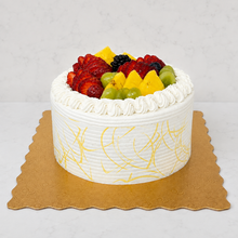Load image into Gallery viewer, A round white cream cake topped with fresh fruit including strawberries, pineapple, grapes, and blackberries, presented on a gold coloured cake board against a light gray marble background.
